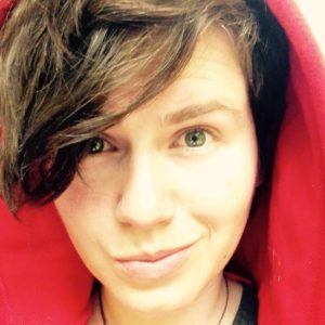 close face shot, libi looks into the camera with a crooked smile. she is white with blue-green eyes and wearing a red hoodie, her brown hair sweeps across her face to the left.